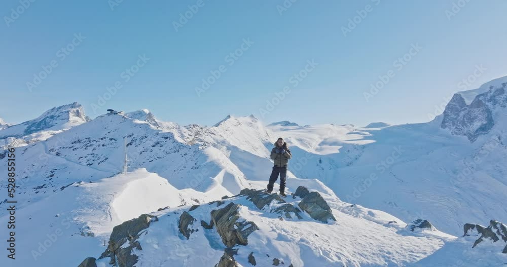 Aerial orbit motion of Young asian man standing on top of the famous Gornergrat observatory operates with drone remote control with iconic famous Matterhorn sunset background. Swiss alps, Switzerland.