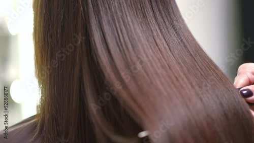 Close-up shaking long laminated hair with hair brush in slow motion. Brunette unrecognizable Caucasian young woman after keratin treatment in beauty parlor indoors with hairstylist photo