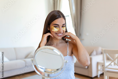 Smiling young woman applying eye hydration mask under her eyes. Female enjoy making face massage, peeling, put on mask under eyes in front of the mirror. Young woman looking in the mirror