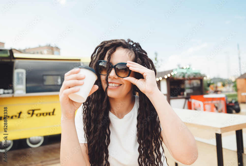 A beautiful cheerful girl with dreadlocks is sitting on a food court and drinking coffee. fast food cafe on the street. a woman drinks coffee in a cafe. autumn