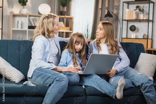 Happy family of mother and two cute daughters sitting together on comfortable couch, using laptop, resting and spend free time at home, recording video in living room. © sofiko14