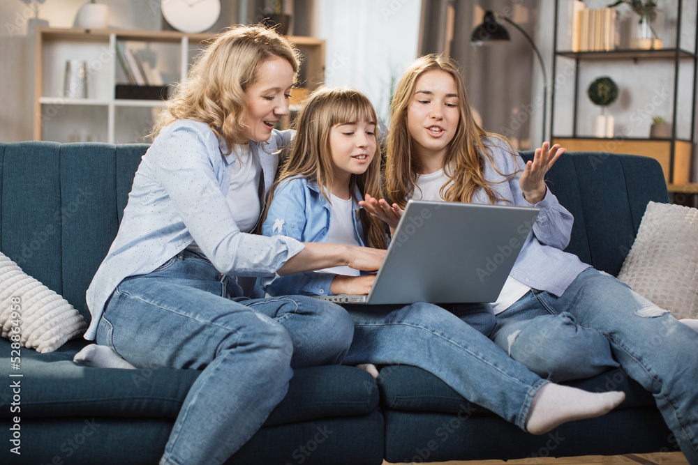 Happy family with two children having good time using laptop. Young smiling mother with cute blond daughters sitting on sofa at home looking at computer screen and watching video. Happy family with two children having good time using laptop. Young smiling mother with cute blond daughters sitting on sofa at home looking at computer screen and watching video.