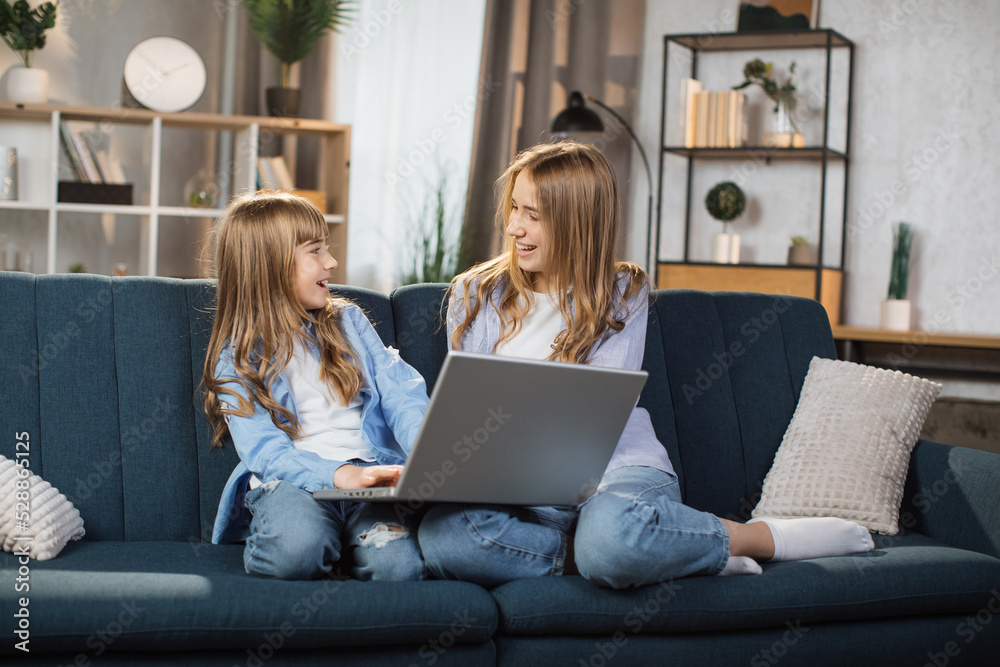 Happy sisters or friends two cute blond girls sitting together on comfortable couch, using laptop, holding video call with father or greeting grandparents, recording video in living room. Happy sisters or friends two cute blond girls sitting together on comfortable couch, using laptop, holding video call with father or greeting grandparents, recording video in living room.