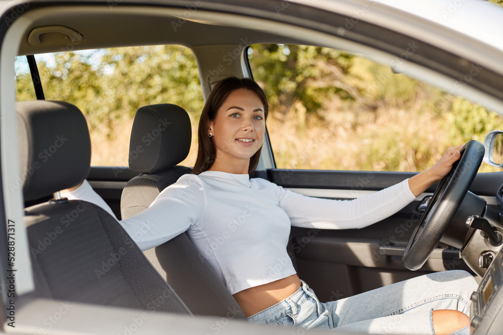 Portrait of smiling self confident dark haired woman wearing white shirt sitting in her car, keeps hand on steering wheel, looking at camera with smile.
