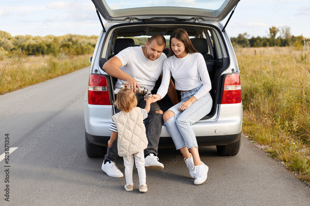 Outdoor shot of loving couple with their baby daughter drinking hot tea from thermos flask sitting in car trunk, stop for resting while spending long hours driving.