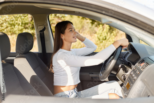 Profile portrait of adorable beautiful young adult Caucasian woman with long dark hair wearing white shirt driving her car, expressing positive emotions. © sementsova321