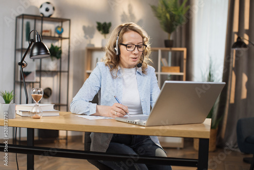 Portrait of beautiful confident with blonde hair and on headphones clever woman wearing casual clothes, sitting at table in front of computer and checking information in presentation on internet