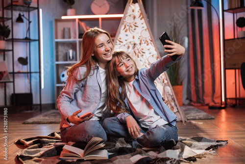 Happy caring nanny or older sister hugging and taking a selfie or receiving a video call with her younger sister during evening time outside teepee tent sitting on the floor at dark cozy room.