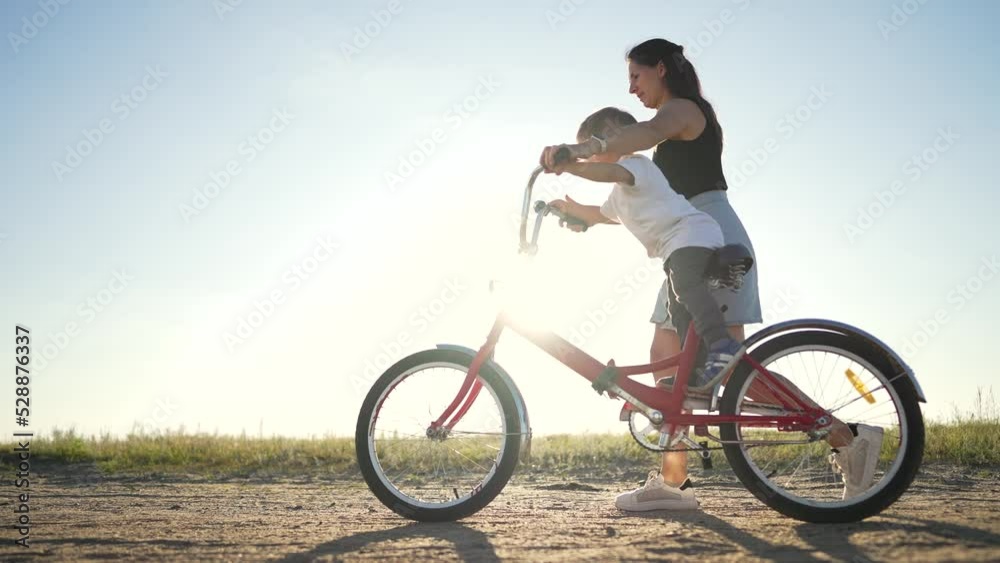Happy family concept. Mom teaches her son to ride bike on green grass. Child rides bicycle along rural road. Green energy. Mom teaches her son to ride bike for the first time in park at sunset. Happy family concept. Mom teaches her son to ride bike on green grass. Child rides bicycle along rural road. Green energy. Mom teaches her son to ride bike for the first time in park at sunset.