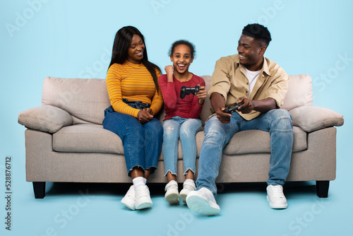 Gaming together. Playful african american father and daughter playing videogames, sitting on sofa over blue background