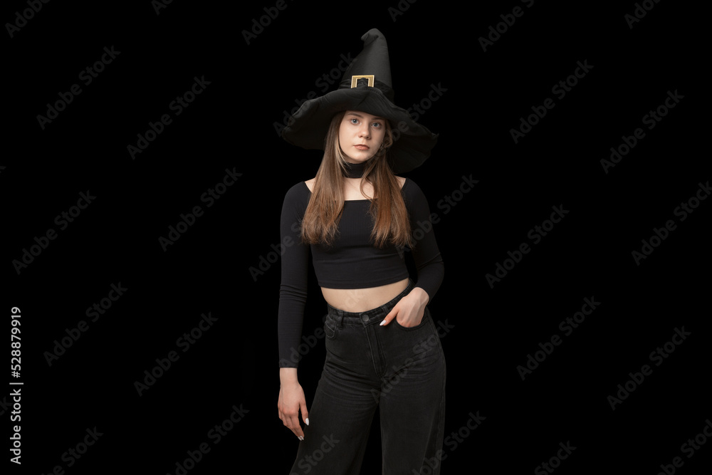 Portrait of young girl in pointed witch hat and black clothes is isolated on black background. Copy space. Halloween costume Portrait of young girl in pointed witch hat and black clothes is isolated on black background. Copy space. Halloween costume