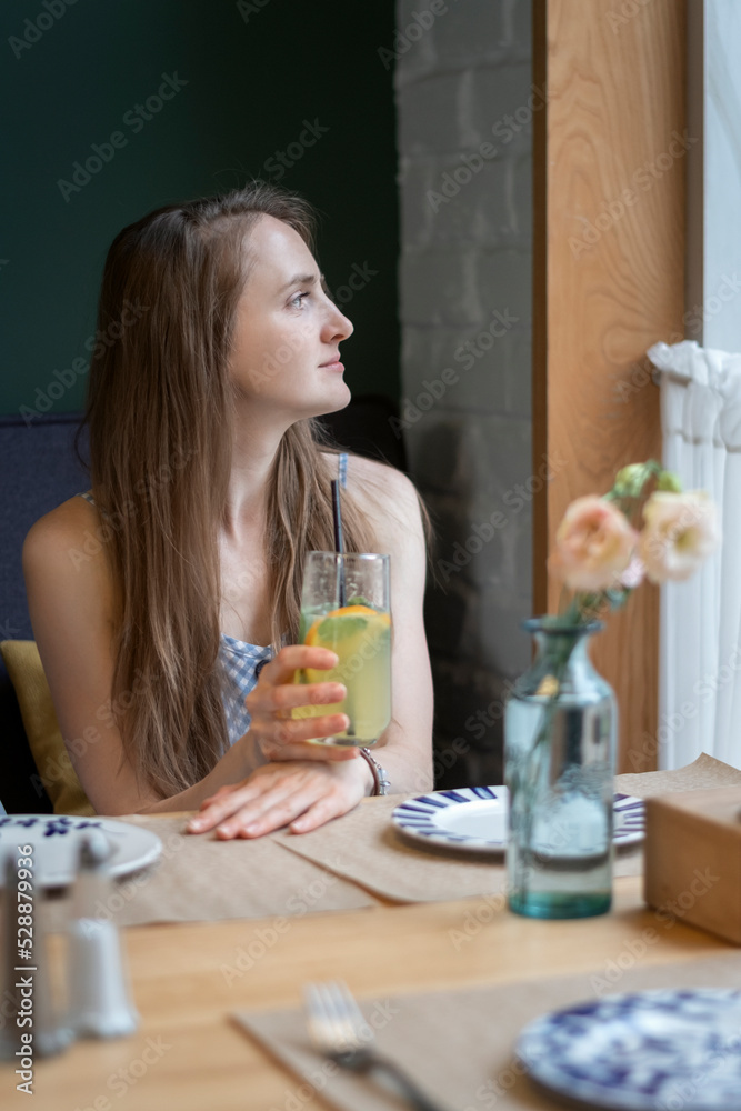 Portrait of young woman holding lemonade or cocktail in hands and looks out the window. Girl at table in cafe waiting for an order. Portrait of young woman holding lemonade or cocktail in hands and looks out the window. Girl at table in cafe waiting for an order.