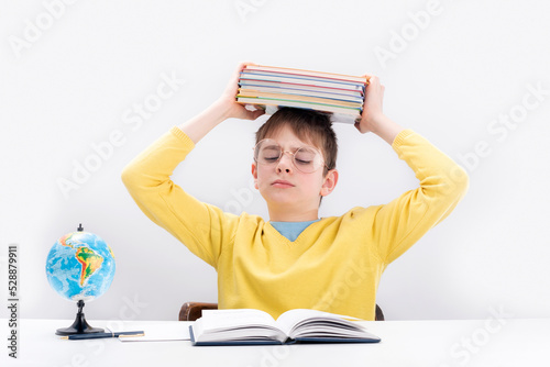 Tired teenage boy sitting at desk and holding stack of notebooks over his head. Schoolboy difficult does his homework