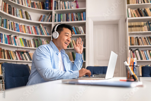 Cheerful Asian Male Waving To Laptop Video Calling In Library