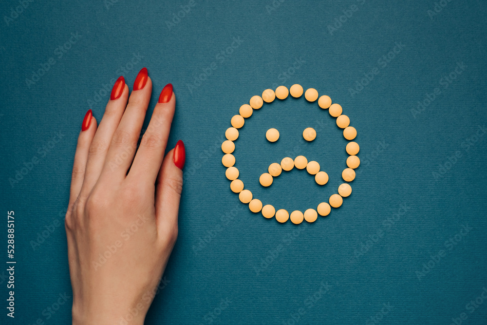 Obraz premium A woman's hand with a red manicure and pills are laid out on a blue background in the form of a sad emoticon. Top view. Antidepressants concept or headache.