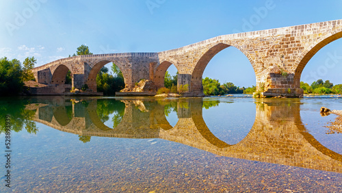 Seljuk Bridge in Aspendos. The Eurymedon Bridge. Crooked bridge, reflected in calm water. An ancient building across the Kopruchay river. Antalya region, Turkey. photo