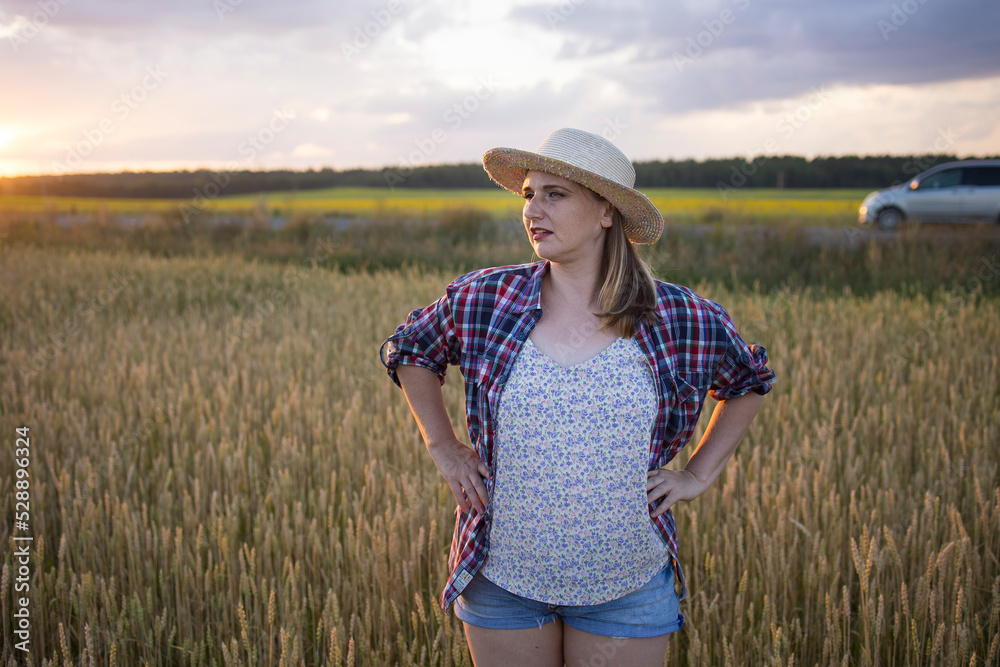 Obraz premium A beautiful middle-aged farmer woman in a straw hat and a plaid shirt stands in a field of golden ripening wheat during the daytime in the sunlight