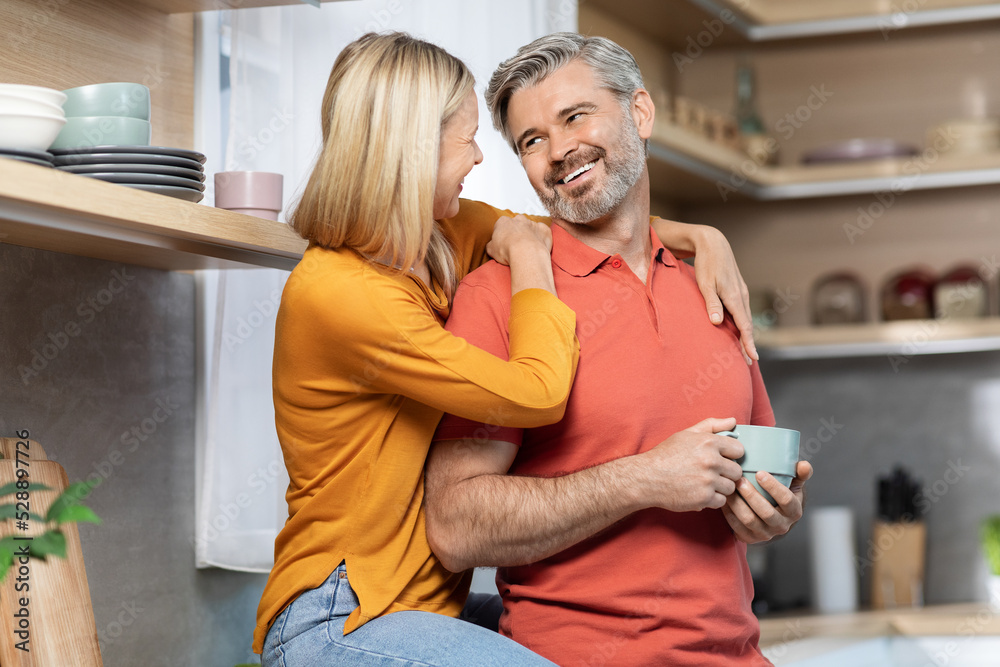 Loving lady hugging her smiling husband, drinking coffee at kitchen Loving lady hugging her smiling husband, drinking coffee at kitchen