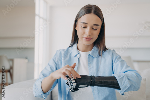 Pretty disabled girl adjusting her bionic prosthetic arm touching sensory panel of artificial hand photo