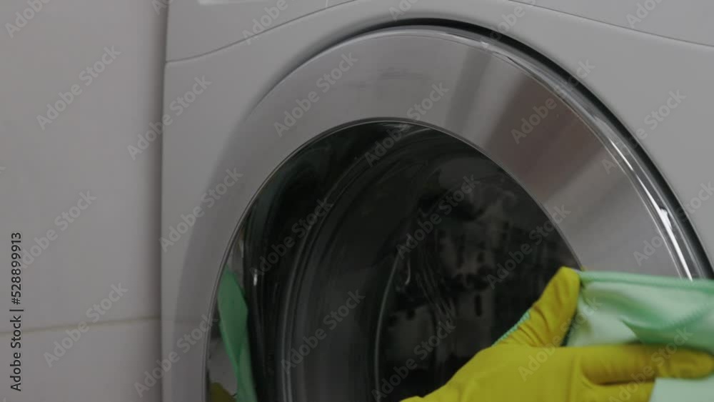 With green napkin with pile girl gently wipes moisture from outside of washing machine door. Carefully to shine woman's gloved hand rubs silver edge of washing machine door while cleaning bathroom. With green napkin with pile girl gently wipes moisture from outside of washing machine door. Carefully to shine woman's gloved hand rubs silver edge of washing machine door while cleaning bathroom.
