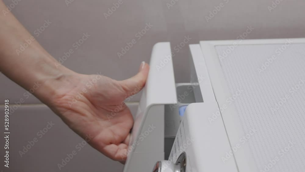 With blue measuring spoon person fills washing machine with powder. Against background of tiles in bathroom girl carefully pours laundry detergent into compartment of washing machine. With blue measuring spoon person fills washing machine with powder. Against background of tiles in bathroom girl carefully pours laundry detergent into compartment of washing machine.