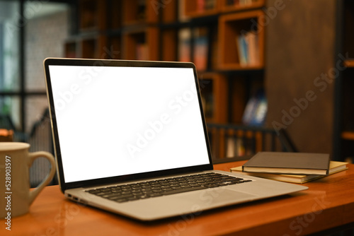 Laptop computer with white empty screen, coffee cup and supplies on wooden table in contemporary office