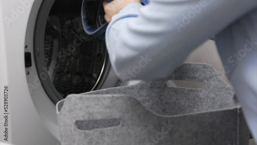 In laundry room, female person loads dirty laundry items into the washing machine. Woman in light colored shirt pulls dirty denim out soft basket and puts them in drum of washing machine for washing. 