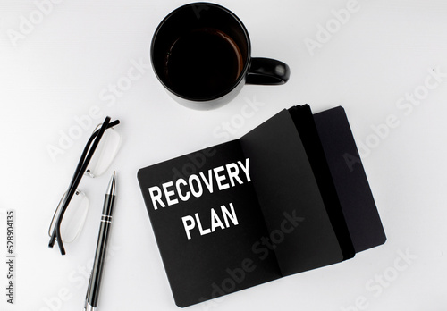 RECOVERY PLAN written text in small black notebook with coffee , pen and glasess on white background. Black-white style
