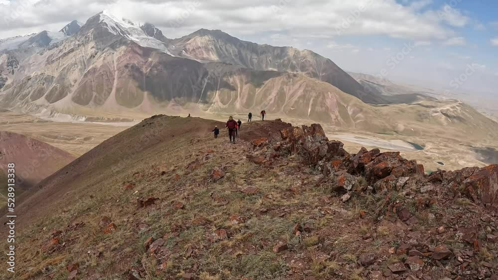 Trekking near the base camp under Lenin Peak, Kyrgyzstan. Several tourists are walking along a mountain path. Beautiful summer mountain landscape. Long shot. Trekking near the base camp under Lenin Peak, Kyrgyzstan. Several tourists are walking along a mountain path. Beautiful summer mountain landscape. Long shot.