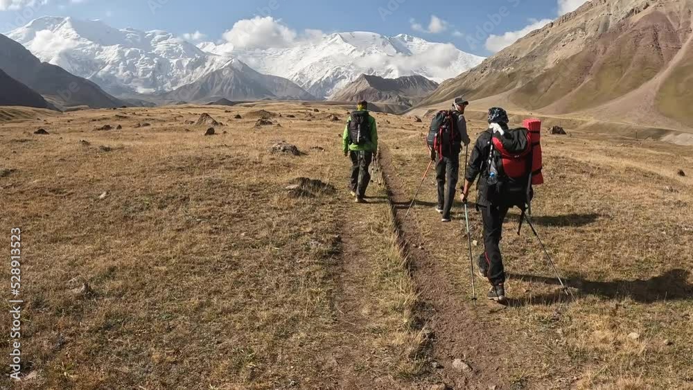 Three tourists with backpacks are walking along the plain along the mountains. Trekking from Base Camp to Camp 1 under Lenin Peak, Kyrgyzstan. Beautiful summer mountain landscape. Three tourists with backpacks are walking along the plain along the mountains. Trekking from Base Camp to Camp 1 under Lenin Peak, Kyrgyzstan. Beautiful summer mountain landscape.