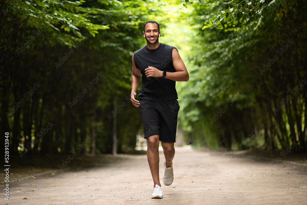 Motivated african american athletic guy jogging by green public park Motivated african american athletic guy jogging by green public park