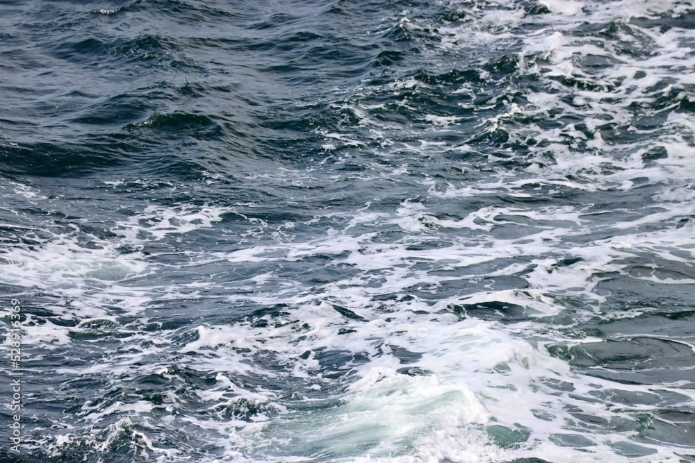 Stern waves with white foam tips on greyish blue sea water, photo taken from aboard ship. Selective focus