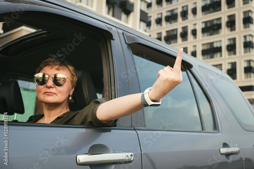 Middle aged woman with sunglasses driving a car showing middle finger. Angry woman with blond hair demonstrating fuck you off sign from open window. © Galina