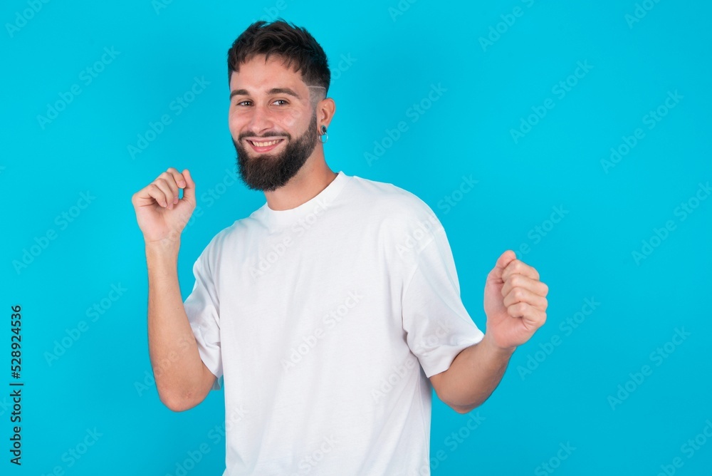 bearded caucasian man wearing white T-shirt over blue background, dancing and listening music with headphones. bearded caucasian man wearing white T-shirt over blue background, dancing and listening music with headphones.