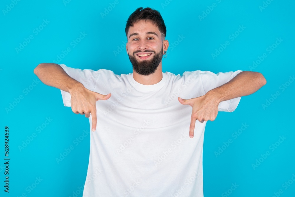 bearded caucasian man wearing white T-shirt over blue background with positive expression, points down with both index fingers, has broad interested smile. Look there, please. bearded caucasian man wearing white T-shirt over blue background with positive expression, points down with both index fingers, has broad interested smile. Look there, please.