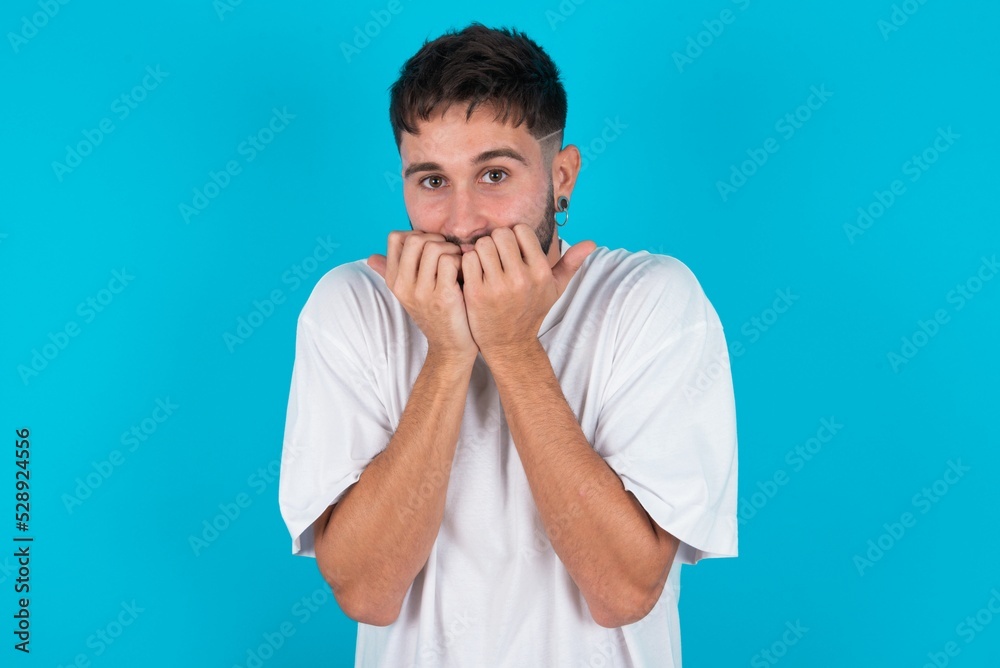 Anxiety - bearded caucasian man wearing white T-shirt over blue background covering his mouth with hands scared from something or someone bitting nails. Anxiety - bearded caucasian man wearing white T-shirt over blue background covering his mouth with hands scared from something or someone bitting nails.