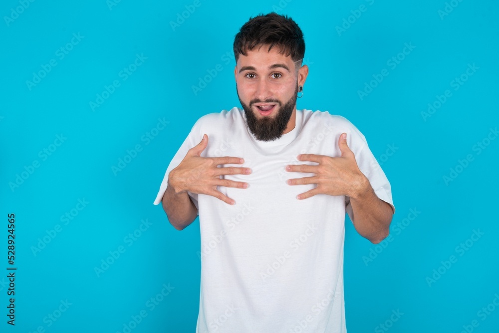 bearded caucasian man wearing white T-shirt over blue background keeps hands on chest feeling shocked and scared, mouth widely opened, stares at camera saying: Who, me? bearded caucasian man wearing white T-shirt over blue background keeps hands on chest feeling shocked and scared, mouth widely opened, stares at camera saying: Who, me?