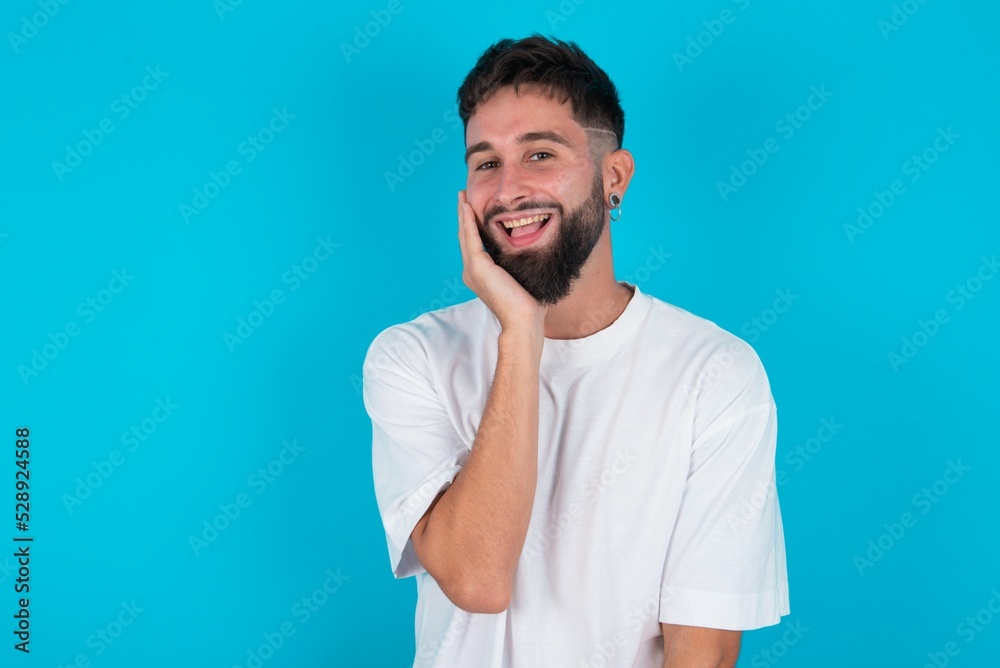 Shocked, astonished bearded caucasian man wearing white T-shirt over blue bac looking surprised in full disbelief wide open mouth with hand near face. Positive emotion facial expression body language.