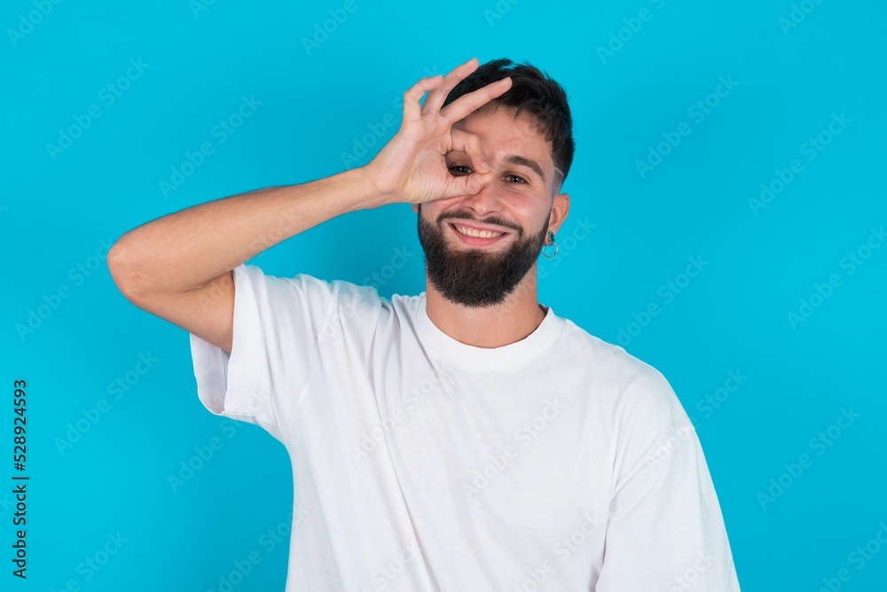 Naklejka premium bearded caucasian man wearing white T-shirt over blue background with happy face smiling doing ok sign with hand on eye looking through finger.