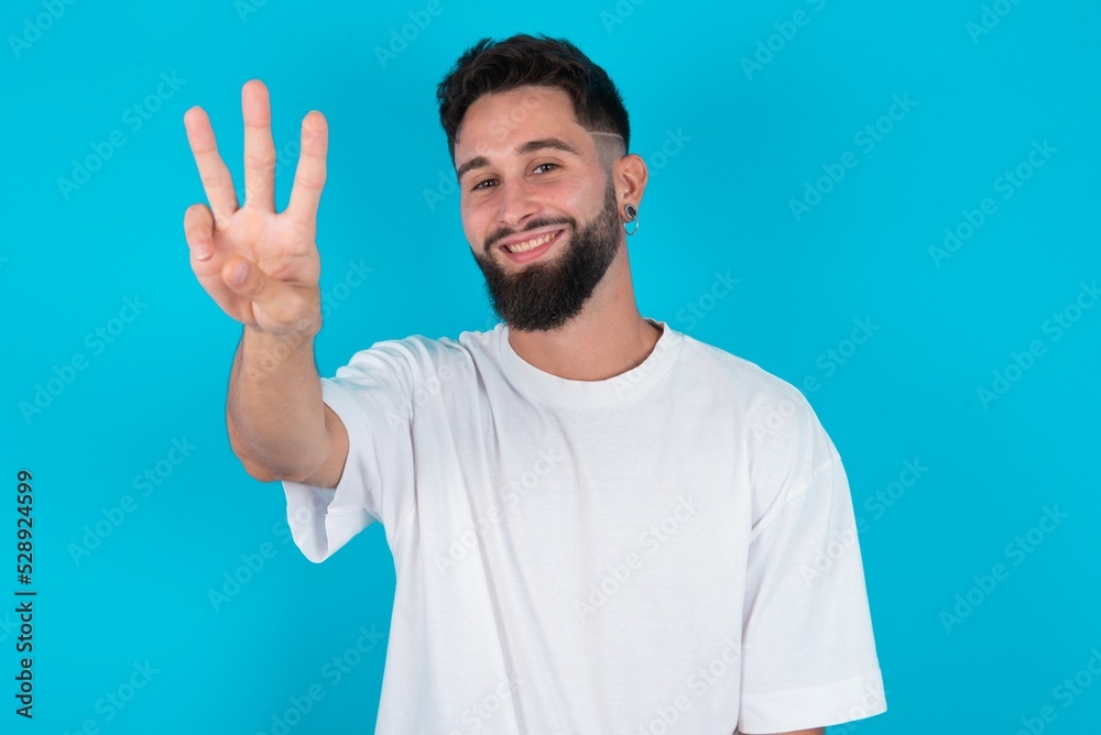 bearded caucasian man wearing white T-shirt over blue background showing and pointing up with fingers number three while smiling confident and happy. bearded caucasian man wearing white T-shirt over blue background showing and pointing up with fingers number three while smiling confident and happy.