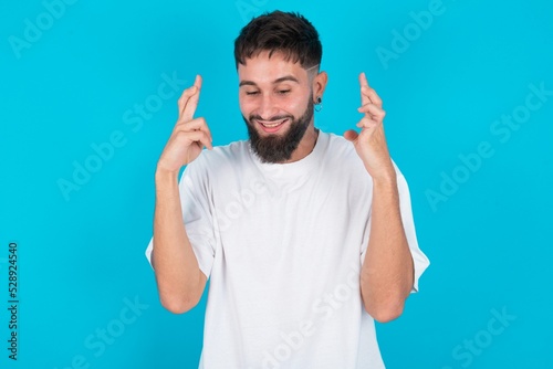 Joyful bearded caucasian man wearing white T-shirt over blue background clenches teeth, raises fingers crossed, makes desirable wish, waits for good news, I have to win.