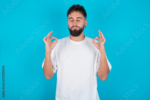 bearded caucasian man wearing white T-shirt over blue background doing yoga, keeping eyes closed, holding fingers in mudra gesture. Meditation, religion and spiritual practices.