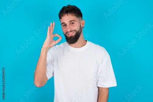 Glad attractive bearded caucasian man wearing white T-shirt over blue background shows ok sign with hand as expresses approval, has cheerful expression, being optimistic.