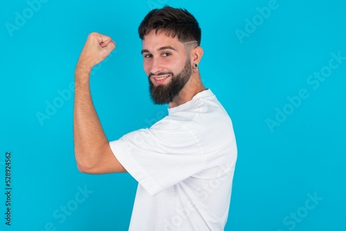 bearded caucasian man wearing white T-shirt over blue background, showing muscles after workout. Health and strength concept.