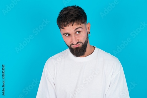 bearded caucasian man wearing white T-shirt over blue background has worried face looking up lips together, being upset thinking about something important, keeps hands down.