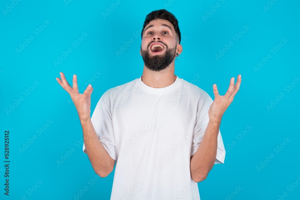 Fototapeta premium Joyful excited lucky bearded caucasian man wearing white T-shirt over blue background cheering, celebrating success, screaming yes with clenched fists