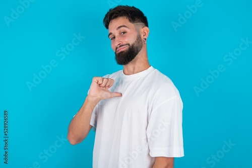 Closeup of cheerful bearded caucasian man wearing white T-shirt over blue background looks joyful, satisfied and confident, points at himself with thumb.