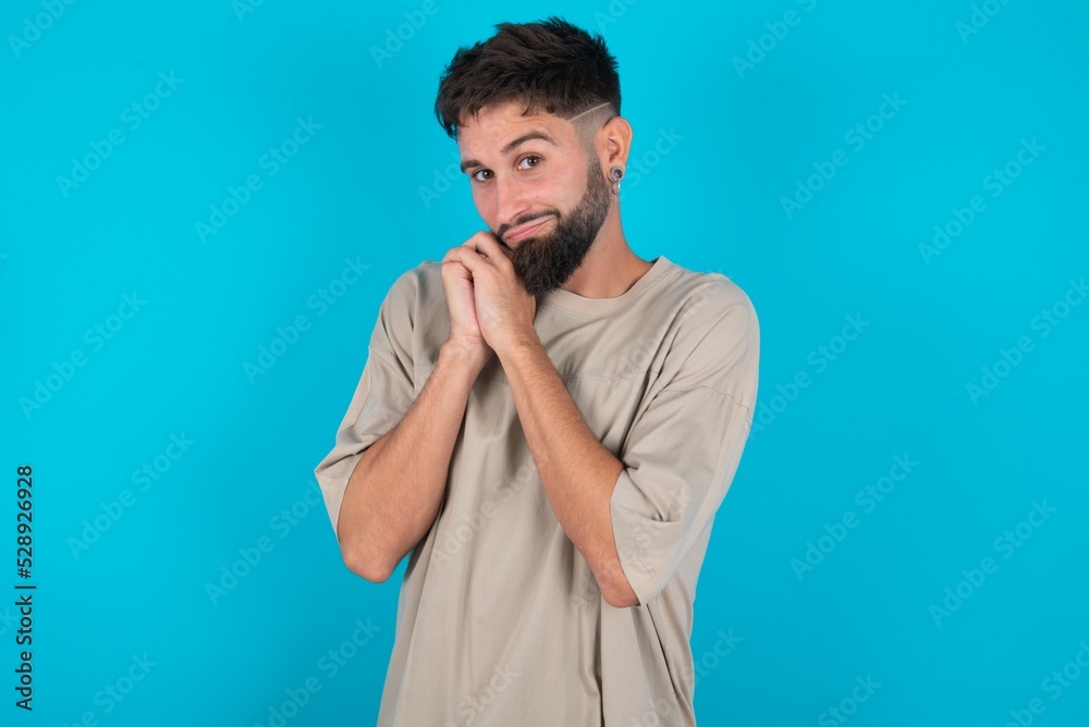 Charming serious bearded caucasian man wearing casual T-shirt over blue background keeps hands near face smiles tenderly at camera Charming serious bearded caucasian man wearing casual T-shirt over blue background keeps hands near face smiles tenderly at camera