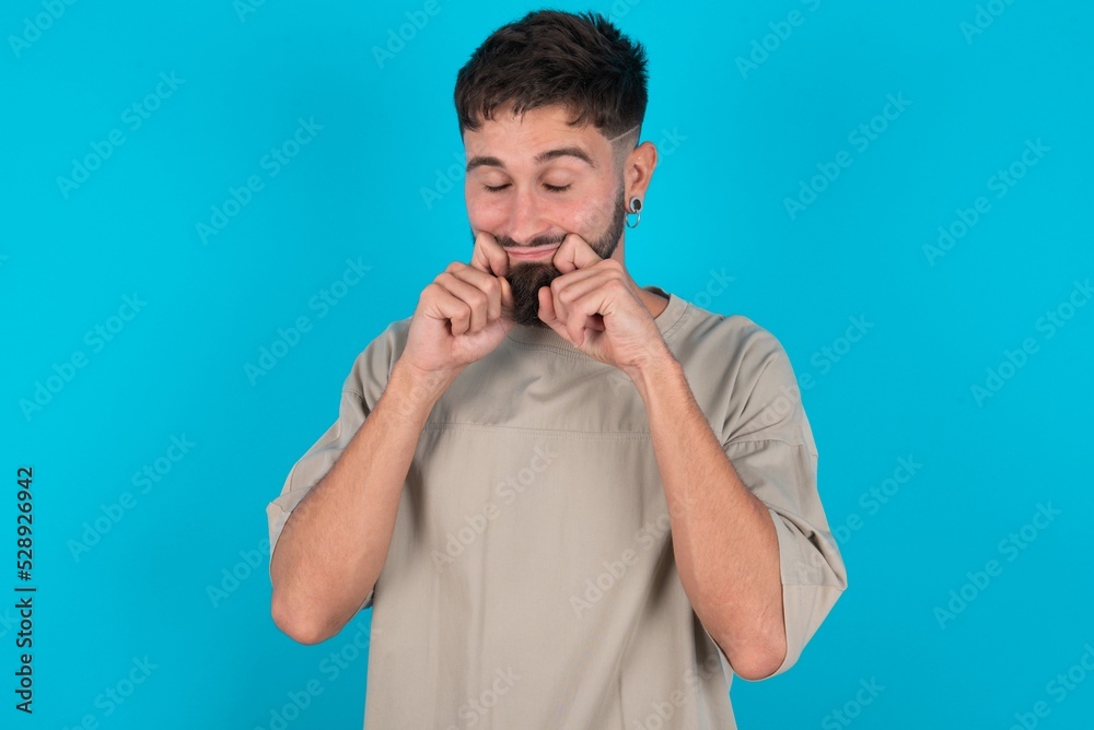 Pleased bearded caucasian man wearing casual T-shirt over blue background with closed eyes keeps hands near cheeks and smiles tenderly imagines something very pleasant Pleased bearded caucasian man wearing casual T-shirt over blue background with closed eyes keeps hands near cheeks and smiles tenderly imagines something very pleasant