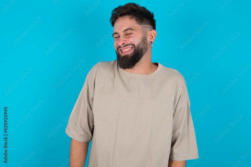 Obraz premium Positive bearded caucasian man wearing casual T-shirt over blue background with overjoyed expression closes eyes and laughs shows white perfect teeth